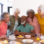 multiracial-senior-friends-with-arms-around-enjoying-by-breakfast-dining-table-nursing-home-cheerful-unaltered-food-drink-healthy-togetherness-support-assisted-living-retirement