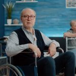Senior man in a wheelchair sitting in a bright living room with another person reading in the background, representing supportive housing and independent living in Vancouver.